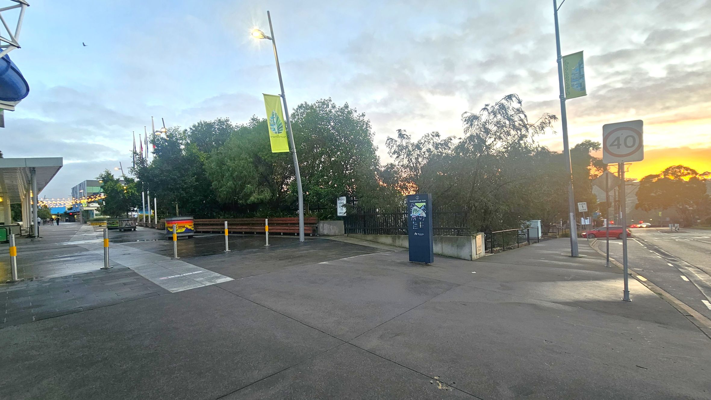 A street scene with a wide pavement in the foreground. On the left, there's a building with a blue awning and a row of flags. In the centre, a path leads towards a cluster of trees and a blue information sign. On the right, a road with a 40 speed limit sign and a red car in the distance. The background features a sunset with orange hues peeking through the trees. The pavement is wet, suggesting recent rain.