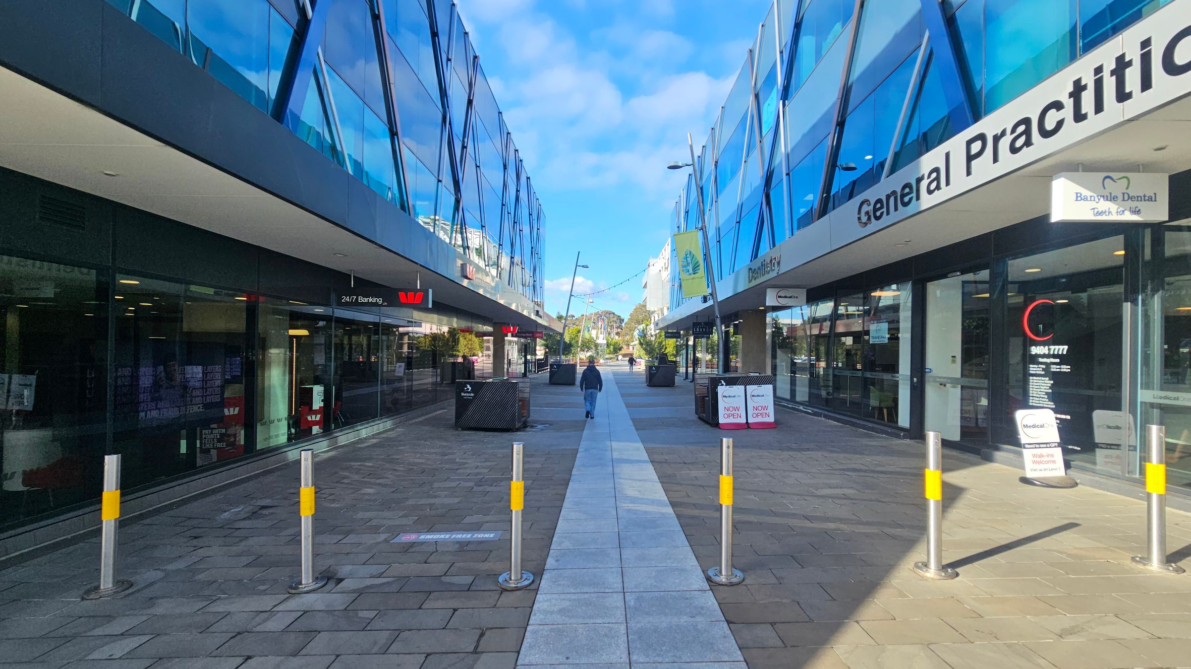 A pedestrian walkway flanked by modern buildings with reflective glass facades. On the left, there's a bank with a "24/7 Banking" sign. On the right, signs for a general practice and dental services are visible. The path is lined with bollards and leads to a distant area with trees. The ground is paved with tiles, and the buildings have overhanging roofs. A person is walking down the centre of the path.