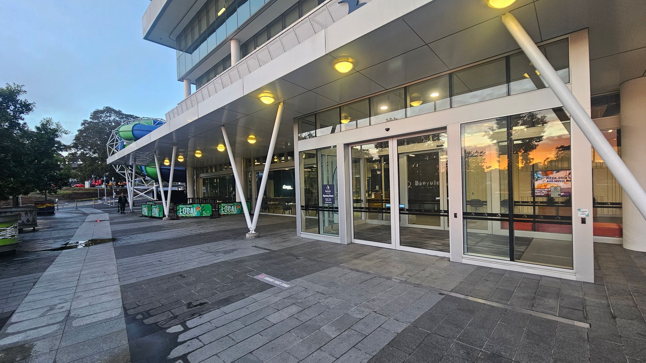 The image shows a modern building with large glass doors and windows on the right. The structure has a white overhang supported by angled pillars, with round lights underneath. On the left, there is a pathway with grey tiles and a few puddles. In the background, a colourful slide structure is visible, along with some trees. A sign on the glass door reads "Banyule." The reflection of a sunset is visible in the glass.