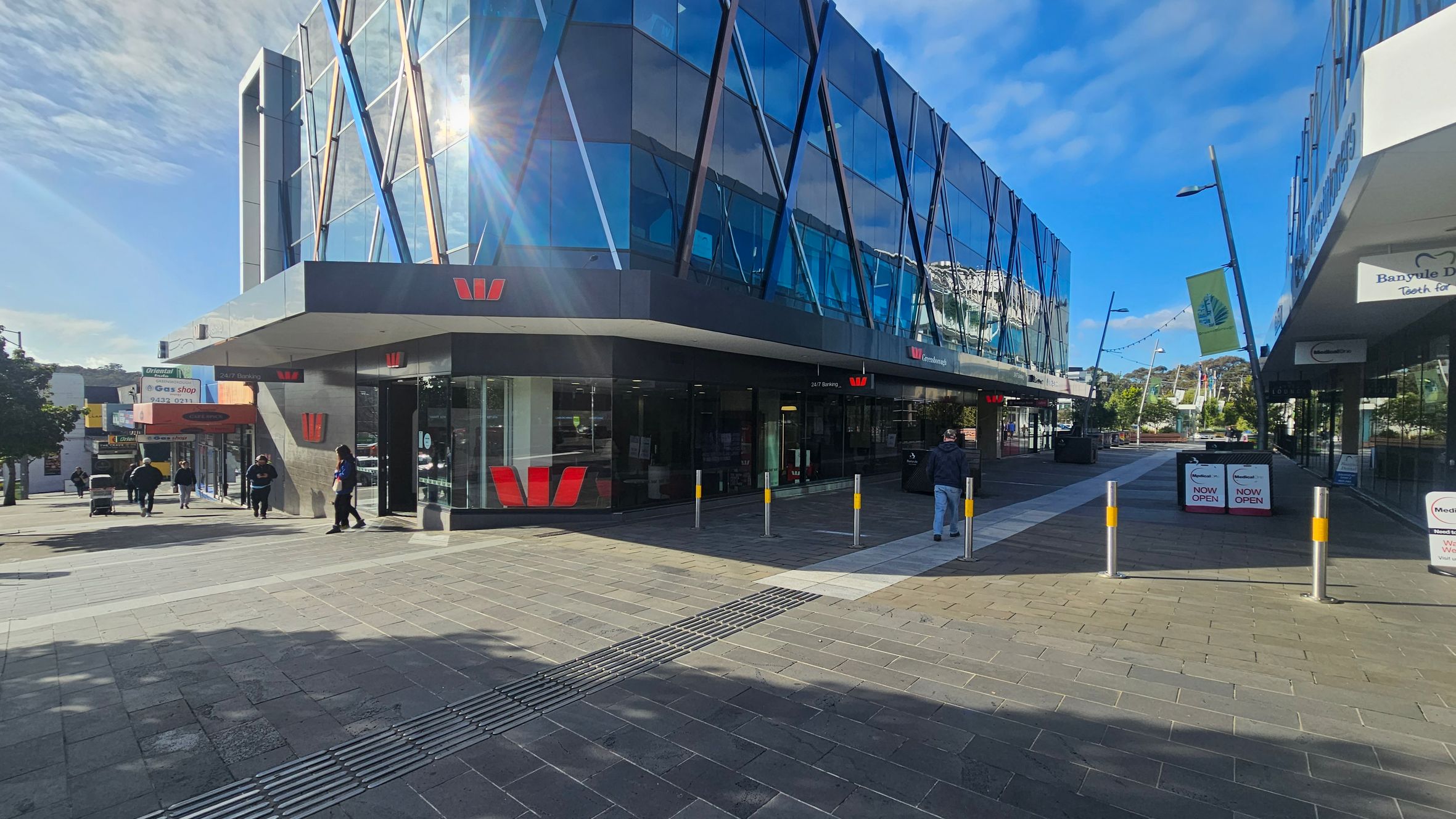 A modern building with a glass facade featuring diagonal blue and white lines is on the left. The building has a prominent red logo. People are walking on the pavement in front of it. On the right, there are more shops with signs and a pathway leading further into the distance. The ground is paved with grey and reddish tiles, and there are several bollards near the entrance. The sky is clear and blue.