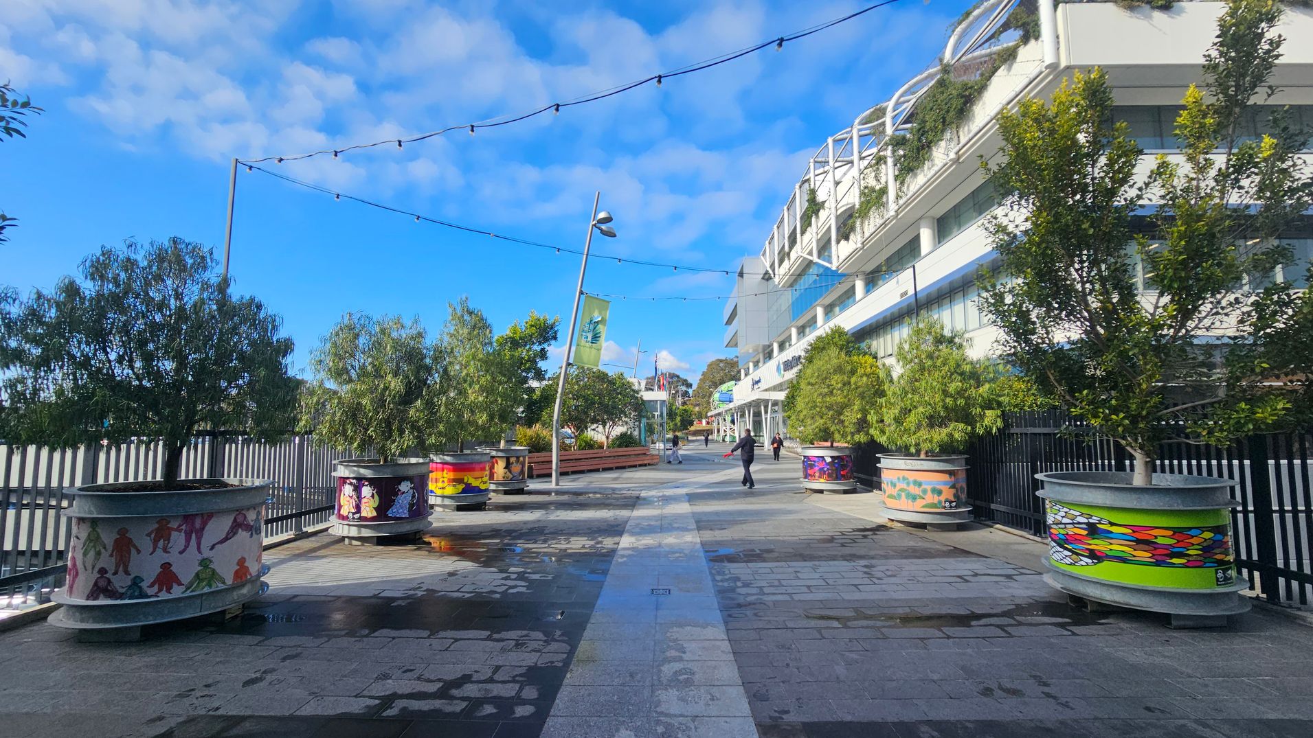 A pedestrian walkway with decorative planters on both sides, each featuring colourful artwork. On the left, trees and a metal fence line the path. On the right, a modern building with glass windows and greenery on the upper levels. Overhead, string lights are suspended between poles. A few people are walking along the path, and a green banner hangs from a pole. The ground is paved with a mix of light and dark tiles, with some wet patches visible.