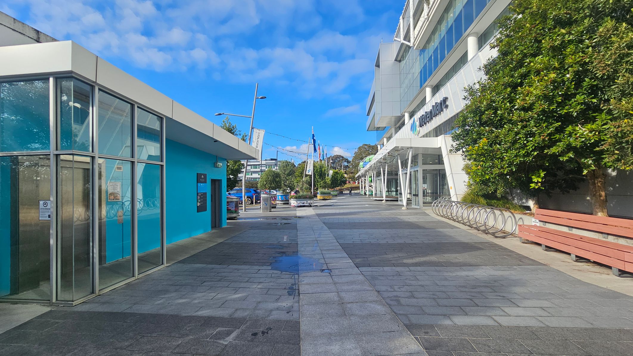 A paved walkway with a building on the left featuring a bright blue wall and large glass windows. On the right, a modern building with a white facade and glass entrance, partially obscured by a tree. A row of bike racks and a wooden bench are visible. In the background, flags and trees line the path, with a few people walking and a blue car parked. The sky is clear with scattered clouds.