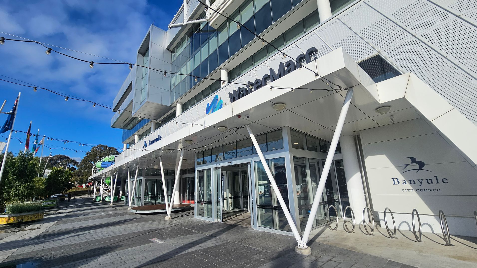 A modern building with a glass facade and angular design elements. The entrance features large glass doors and a sign reading "WaterMarc" above. To the right, there's a "Banyule City Council" sign. Several white poles support the structure, and string lights are suspended above. Flags are visible on the left, and there are trees and planters along the paved walkway. The sky is clear and blue.