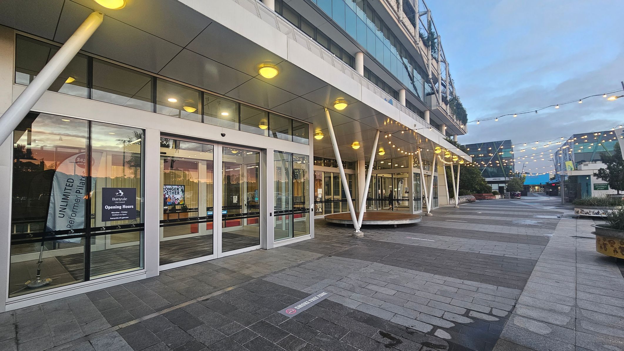 A modern building entrance with large glass doors on the left, displaying signs and banners. The ceiling features round lights, and the floor is paved with grey tiles. White structural beams support the overhang. To the right, a pedestrian walkway is adorned with string lights overhead. The area is open, with planters and benches along the path. The background shows more buildings with glass facades.