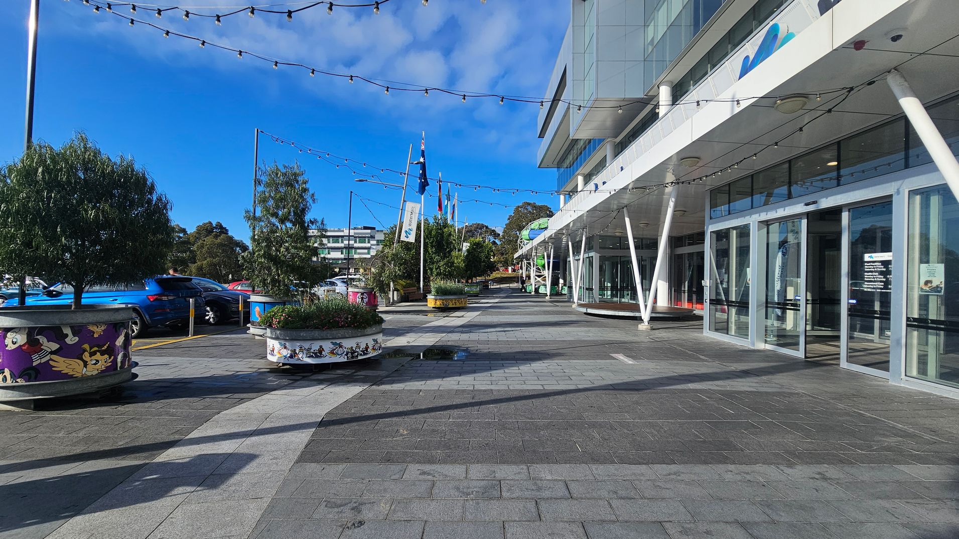A modern building with large glass doors and a white overhang supported by angled pillars is on the right. The pavement is grey with a lighter path running through it. On the left, there are decorative planters with cartoon designs, containing small trees. Several cars are parked nearby. Flags are visible in the background, and string lights are hung above the area.