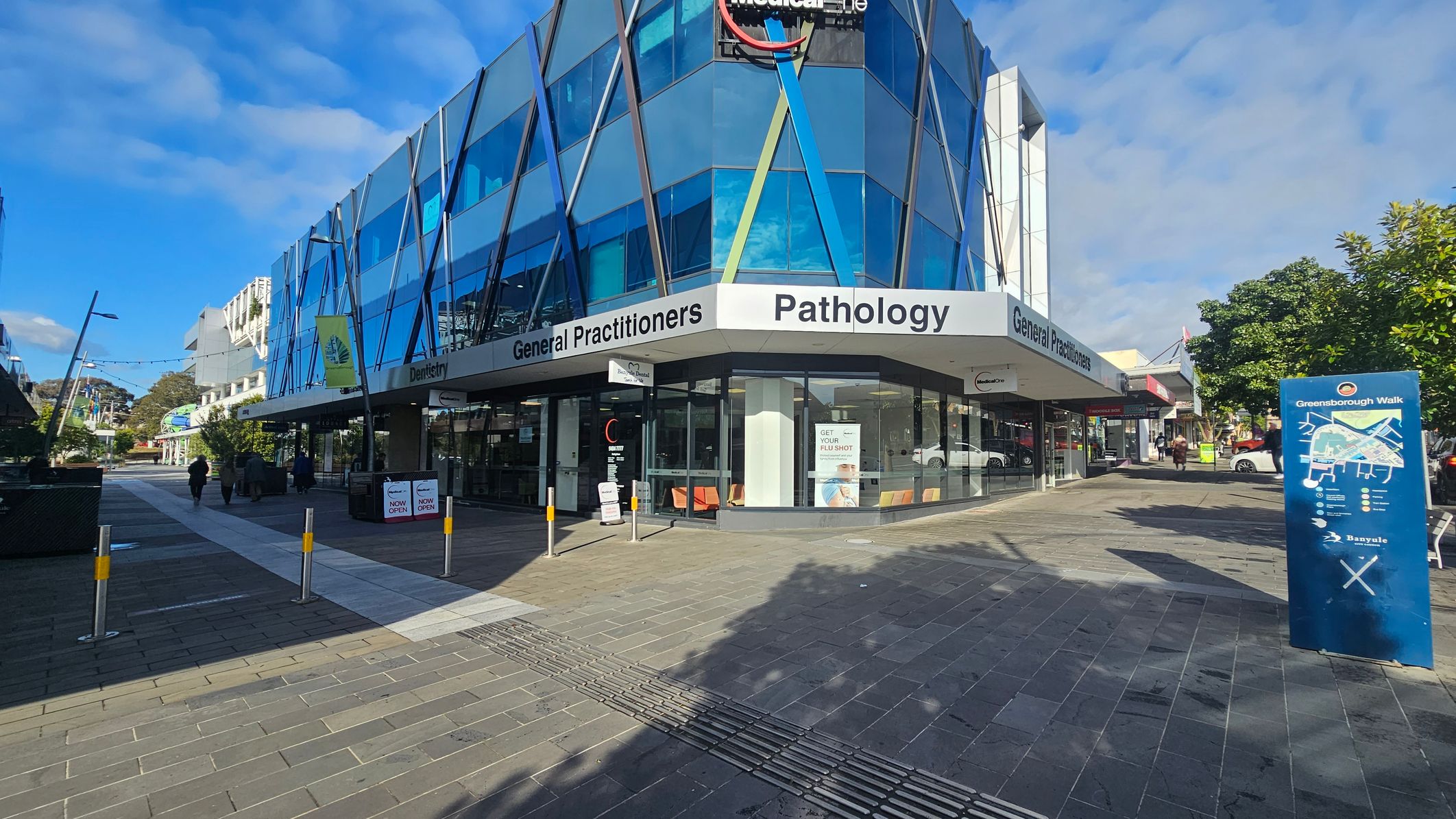 A modern building with blue-tinted glass panels and angular design is on the corner. Signs for "General Practitioners" and "Pathology" are visible. The pavement is a mix of grey and red tiles. On the right, a blue directory sign reads "Greensborough Walk". Trees and a few pedestrians are in the background. The area is well-lit with clear skies.