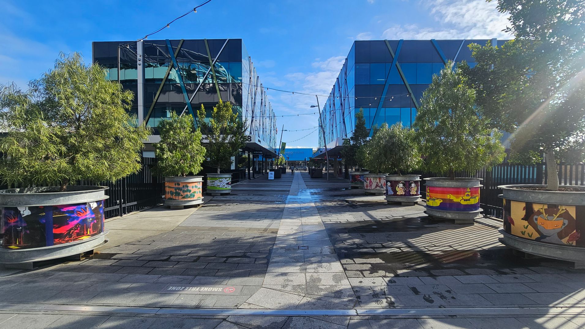 A pedestrian walkway flanked by two modern glass buildings with crisscrossing metal beams. Large, round planters with various colourful designs line both sides of the path, each containing a small tree. Overhead, string lights are suspended between the buildings. The ground is paved with grey tiles, and there are shadows cast by the trees and planters. In the distance, a blue sign is visible at the end of the walkway.