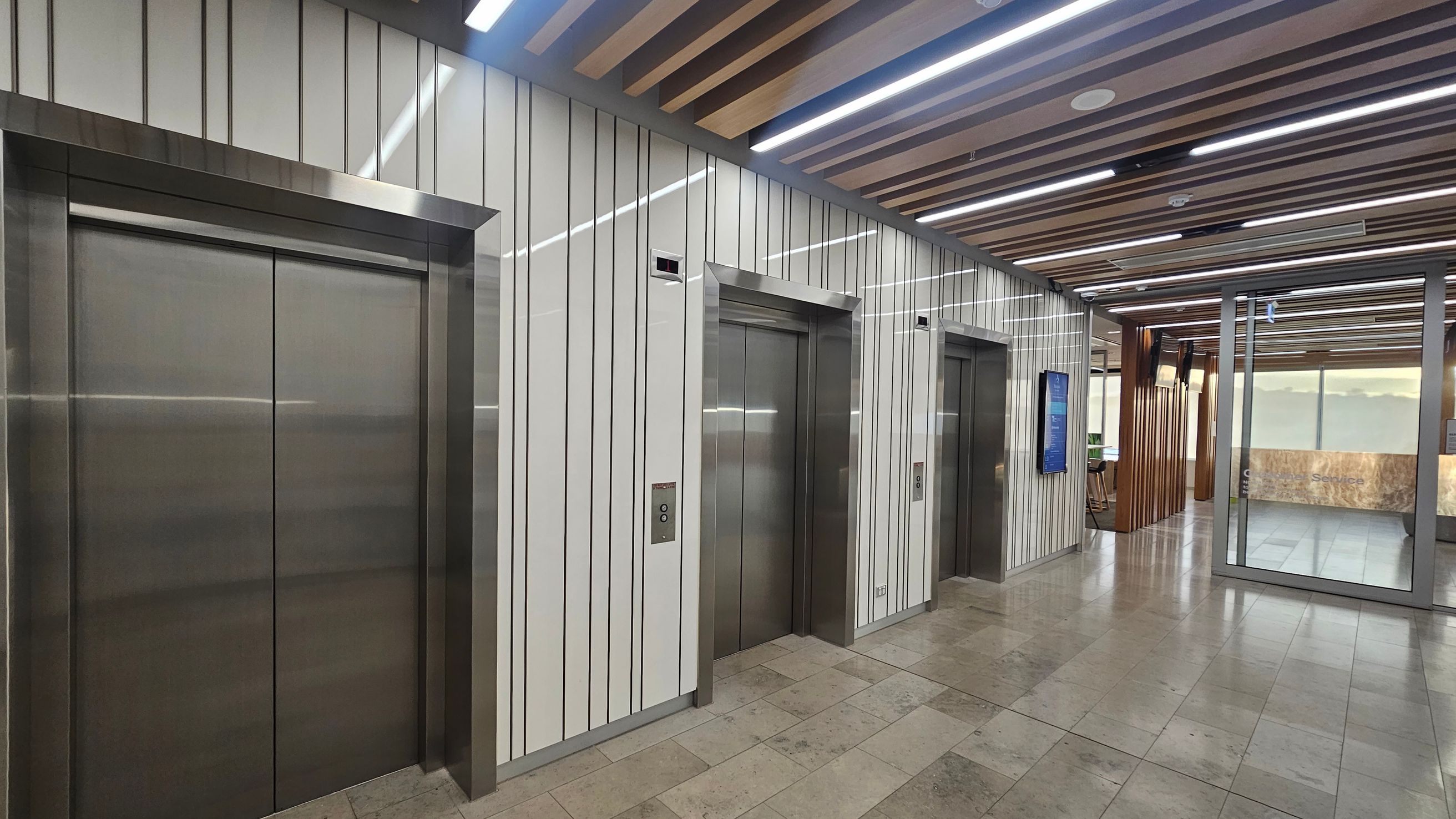 Three lift doors are aligned on the left, set within a wall with vertical striped panels. Each lift has a metallic frame and a button panel beside it. The floor is tiled in a mix of light and dark shades. The ceiling features wooden slats and linear lights. On the right, a glass partition leads to another area with a reception desk and seating. A digital display is mounted on the wall near the lifts.