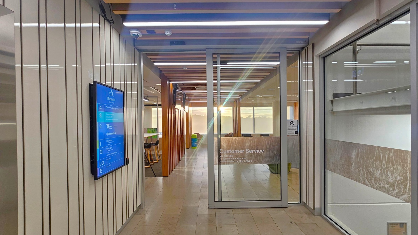 A modern office hallway with a light wooden floor and a ceiling featuring wooden slats and recessed lighting. On the left, a digital directory is mounted on a wall with vertical lines. To the right, a glass door marked "Customer Service" leads to a room with frosted glass panels. In the background, there are high stools and tables, with sunlight streaming through large windows, illuminating the space.
