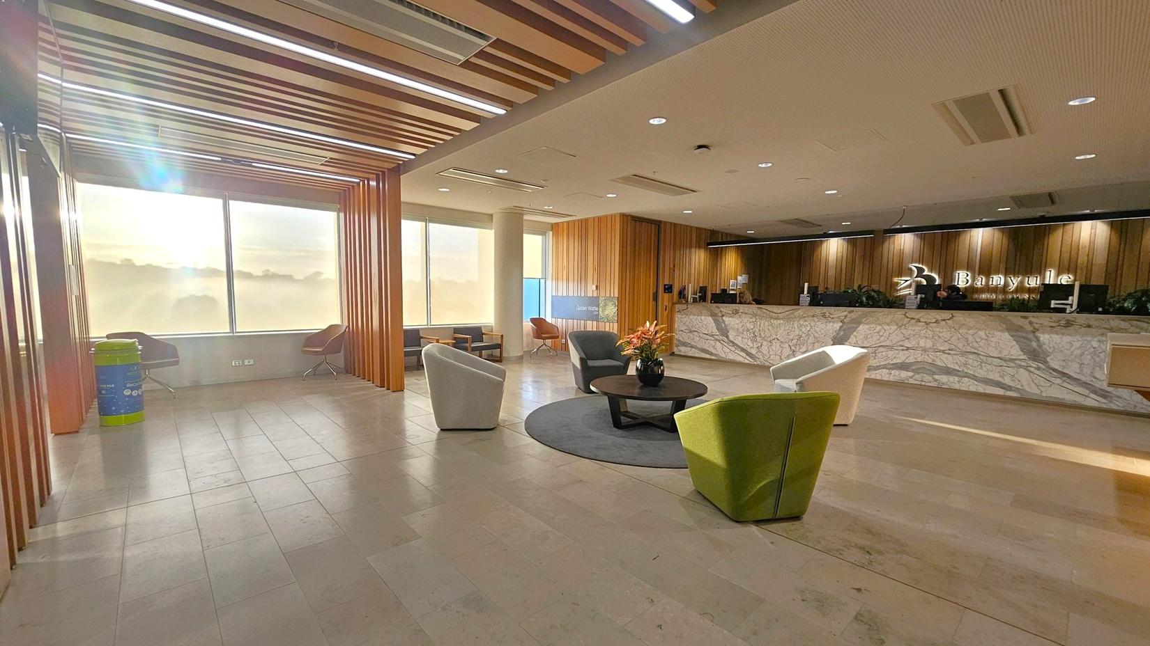 A spacious lobby with a marble reception desk on the right, featuring the name "Banyule" on the wall behind it. In the centre, there is a seating area with a round table and chairs in various colours, including green and white. Large windows on the left allow natural light to flood in, with a recycling bin and additional seating near the windows. The ceiling has wooden slats and recessed lighting, and the floor is tiled in a light stone pattern.