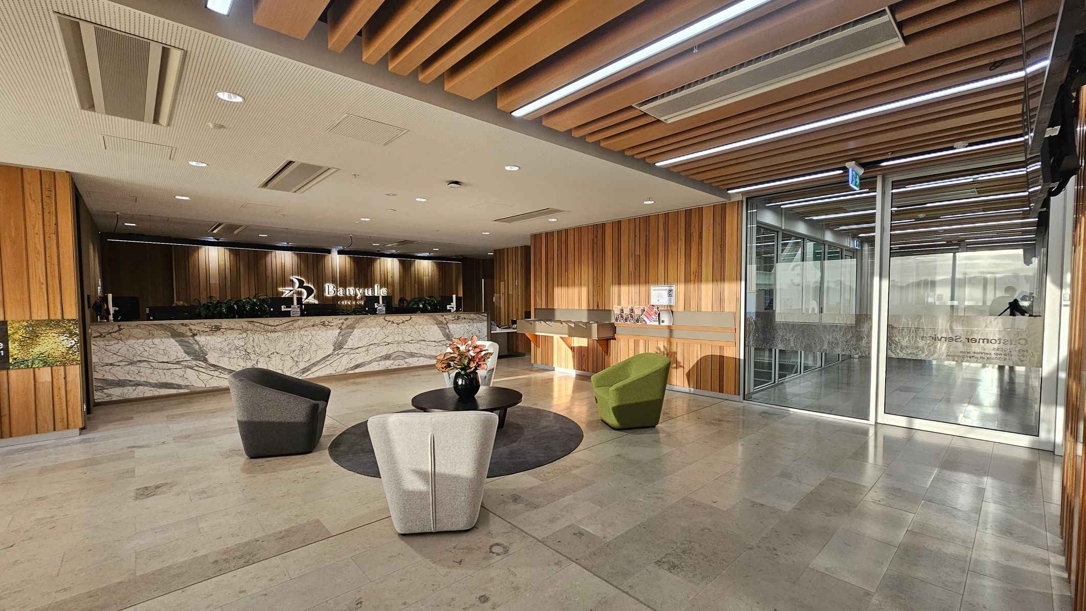 A modern reception area with a marble desk on the left, featuring a logo and plants. In the centre, there are three upholstered chairs around a circular table with a vase of flowers. The right side has glass doors with signage and a wooden wall with brochures. The ceiling has wooden slats and recessed lighting, while the floor is tiled in a light stone pattern.