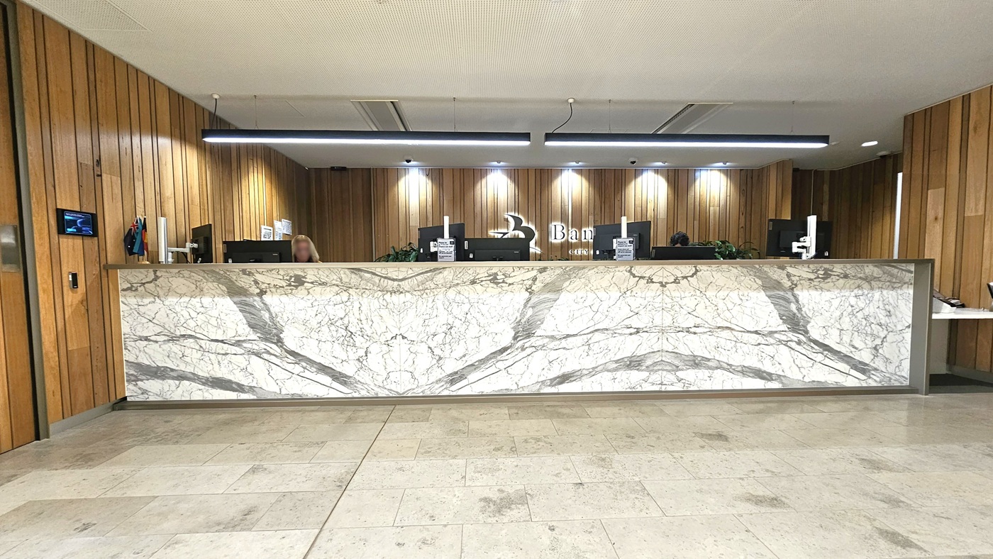 A reception area with a large marble desk featuring grey and white veining. Behind the desk, there are several computer monitors and a person working. The background has wooden panelling with a logo and the word "Bank" visible. The ceiling has recessed lighting and a modern light fixture above the desk. The floor is tiled in a light stone pattern. On the left wall, there is a small screen and a couple of small flags.
