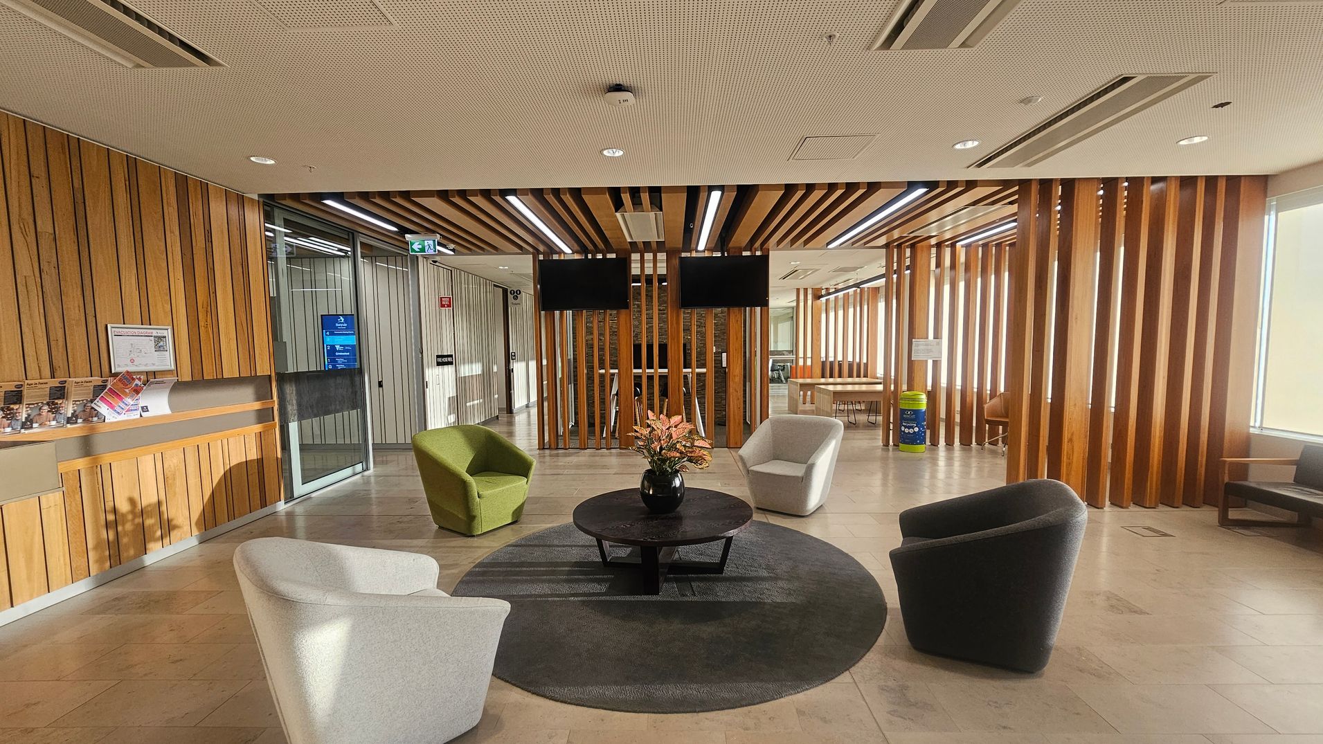 A modern waiting area with wooden slat walls and ceiling. In the centre, a round table with a vase of flowers is surrounded by four cushioned chairs in green, grey, and white. A circular rug lies beneath the table. On the left, a counter with brochures and a glass door. Two black screens are mounted on the wooden partition. On the right, a recycling bin and a window with blinds. The floor is tiled, and the ceiling has recessed lighting and air vents.
