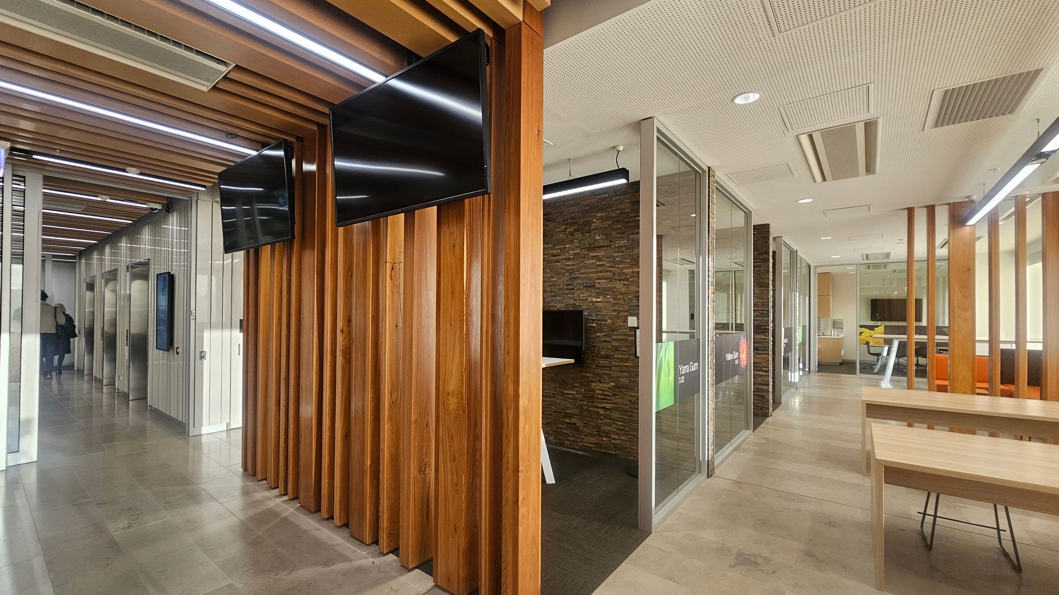 A modern office interior with a corridor on the left featuring elevators and a person walking away. The ceiling has wooden slats with integrated lighting. On the right, there are glass-walled meeting rooms with stone-textured walls and wooden partitions. A wooden table with chairs is in the foreground. Two black screens are mounted on the wooden slats. The floor is tiled, and the ceiling has recessed lighting and ventilation panels.
