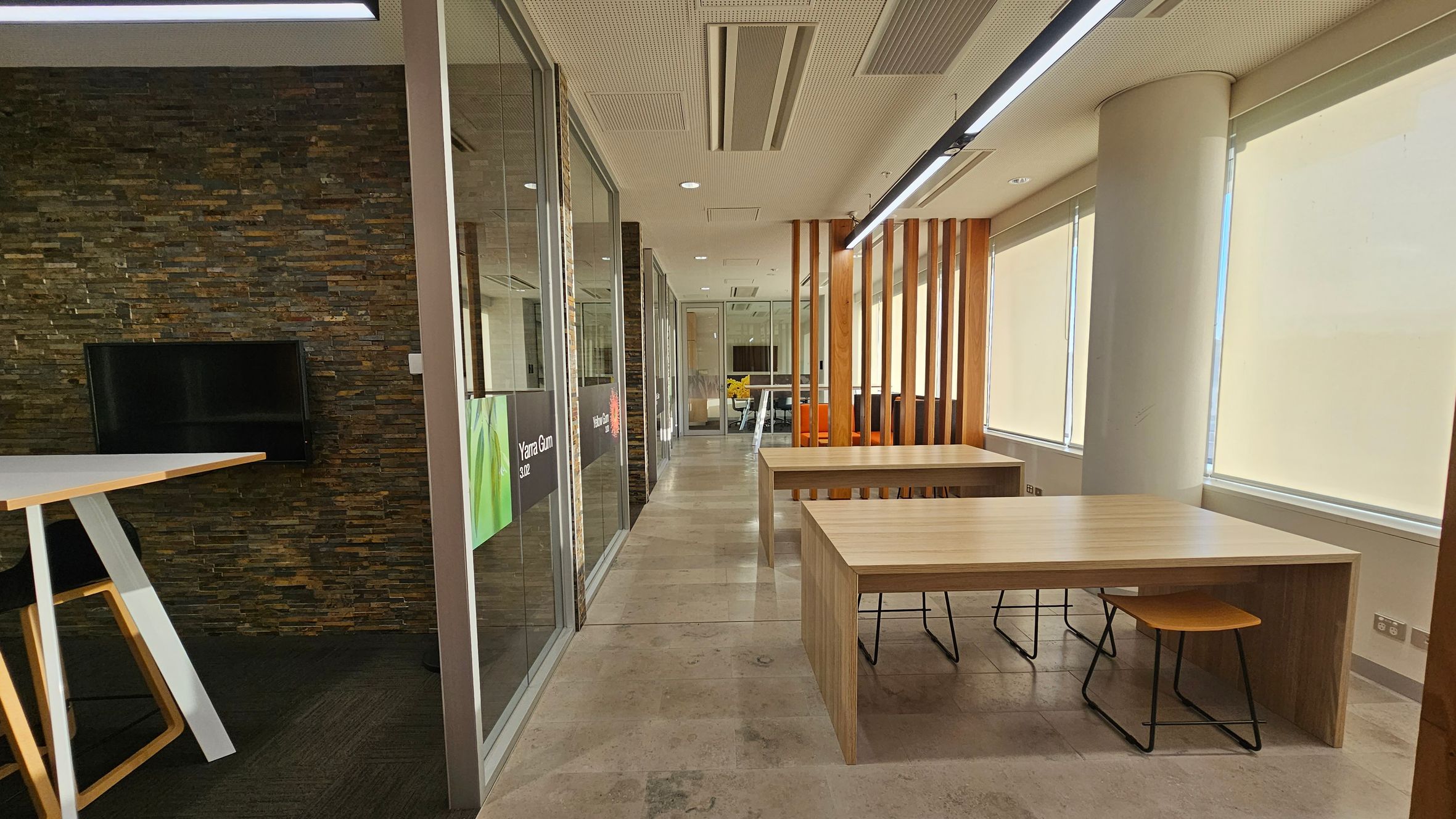 A modern office space with a stone-textured wall on the left featuring a mounted TV. A high table with a stool is nearby. Glass partitions with signage separate the area. On the right, large windows with blinds allow natural light in, and there are wooden tables with stools. A wooden slat partition divides the space. The floor is tiled, and the ceiling has recessed lighting and air vents.
