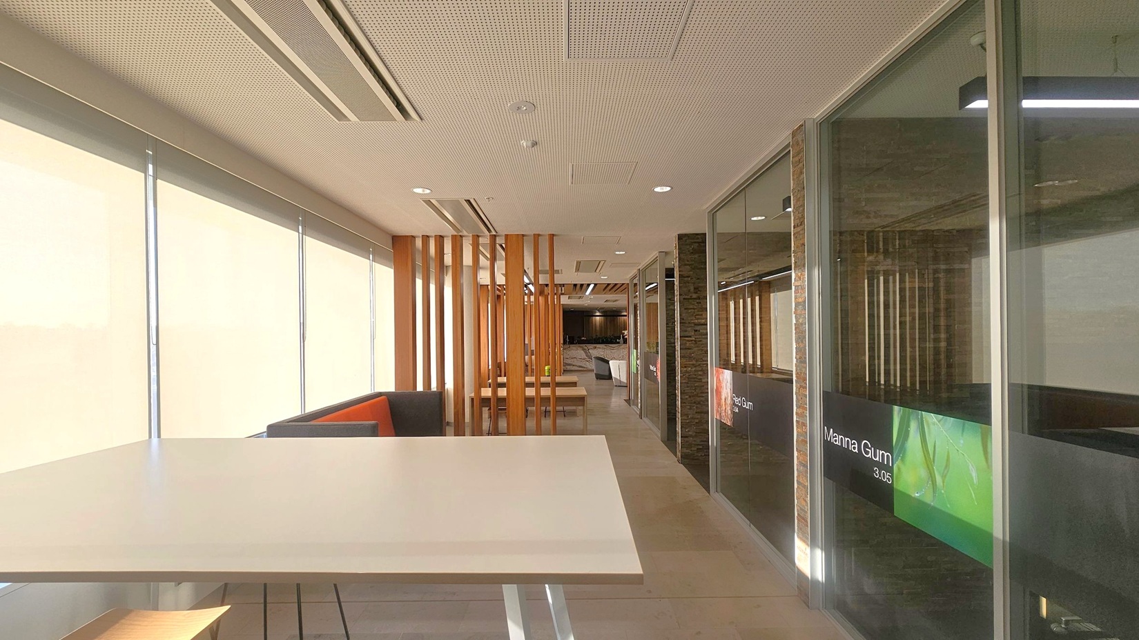 A modern office corridor with a light-coloured ceiling featuring recessed lighting and air vents. On the left, large windows with blinds allow natural light to filter in. Below the windows, a long table with a light surface is visible. On the right, glass walls with signage, including "Manna Gum 3.05," separate the corridor from adjacent rooms. Wooden slats and seating areas are visible further down the corridor, with a tiled floor running throughout.