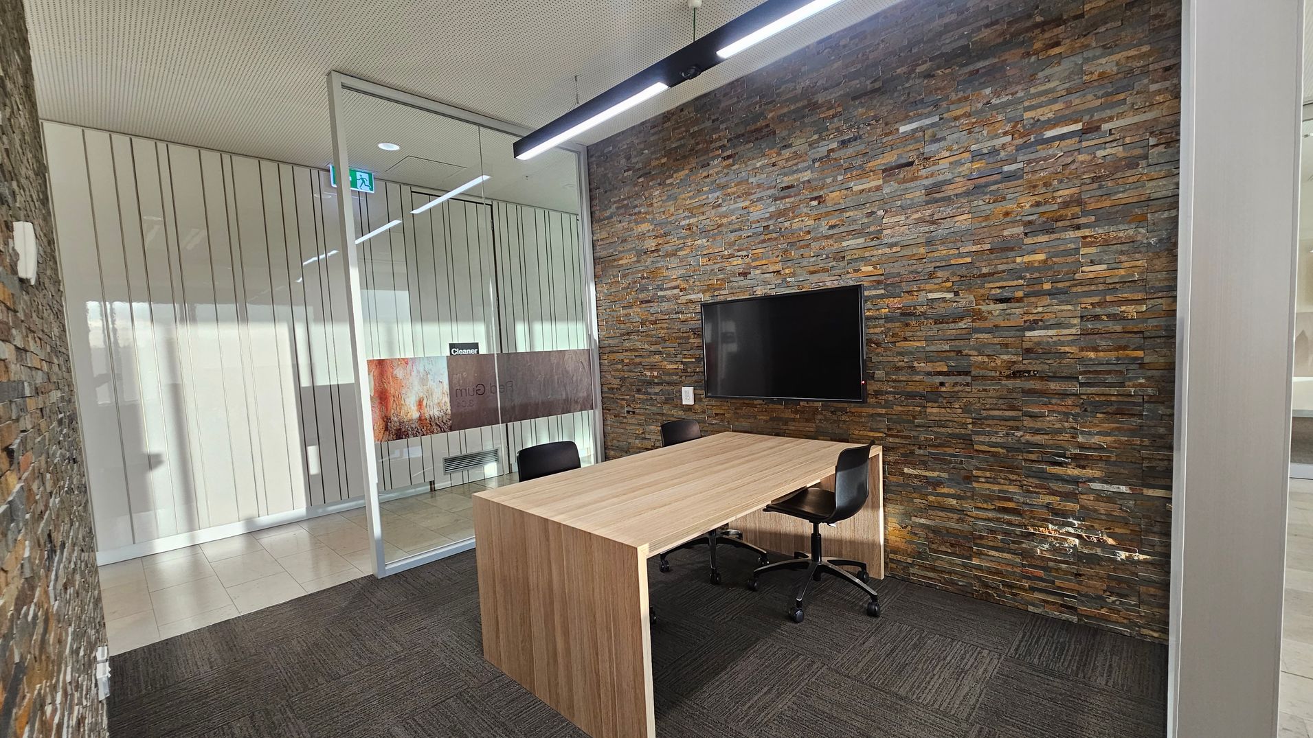 A modern office space with a textured stone wall on the right, featuring a mounted flat-screen TV. In the centre, a wooden table with two black swivel chairs. The left side has a glass partition with vertical lines, leading to another room. The floor is covered with dark grey carpet tiles, and the ceiling is light-coloured with a linear light fixture.