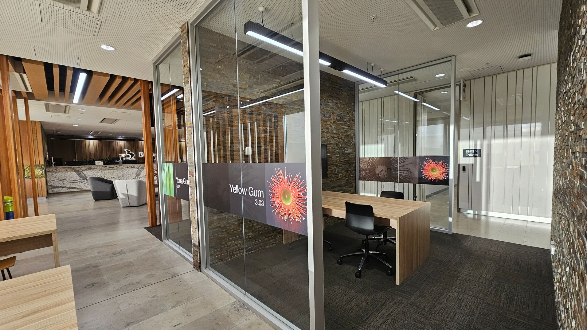 A modern office space with a glass-walled meeting room on the right, labelled "Yellow Gum 3.03," featuring a wooden table and black chairs. The background shows a reception area with a marble counter and grey chairs. The ceiling has recessed lighting and wooden slats. The floor is a mix of light wood and dark carpet. A sign for toilets is visible on the right wall.