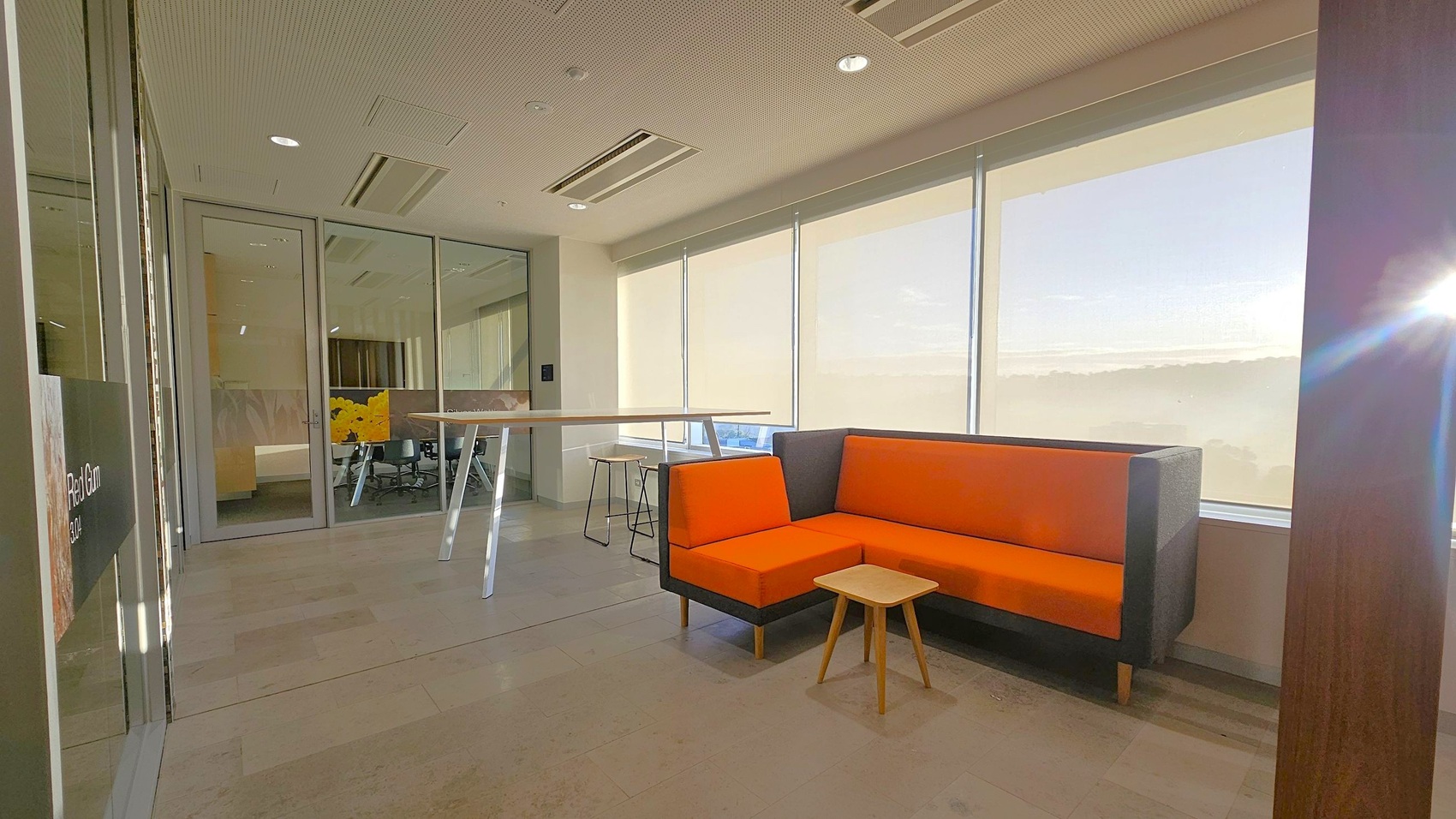 A modern office space with a small seating area on the right, featuring an orange and grey corner sofa and a small wooden table. Large windows with blinds allow natural light to fill the room. On the left, a glass wall separates the seating area from a meeting room, which contains chairs and a table. The floor is tiled, and the ceiling has recessed lighting and air conditioning vents. A high table with stools is positioned near the glass wall.