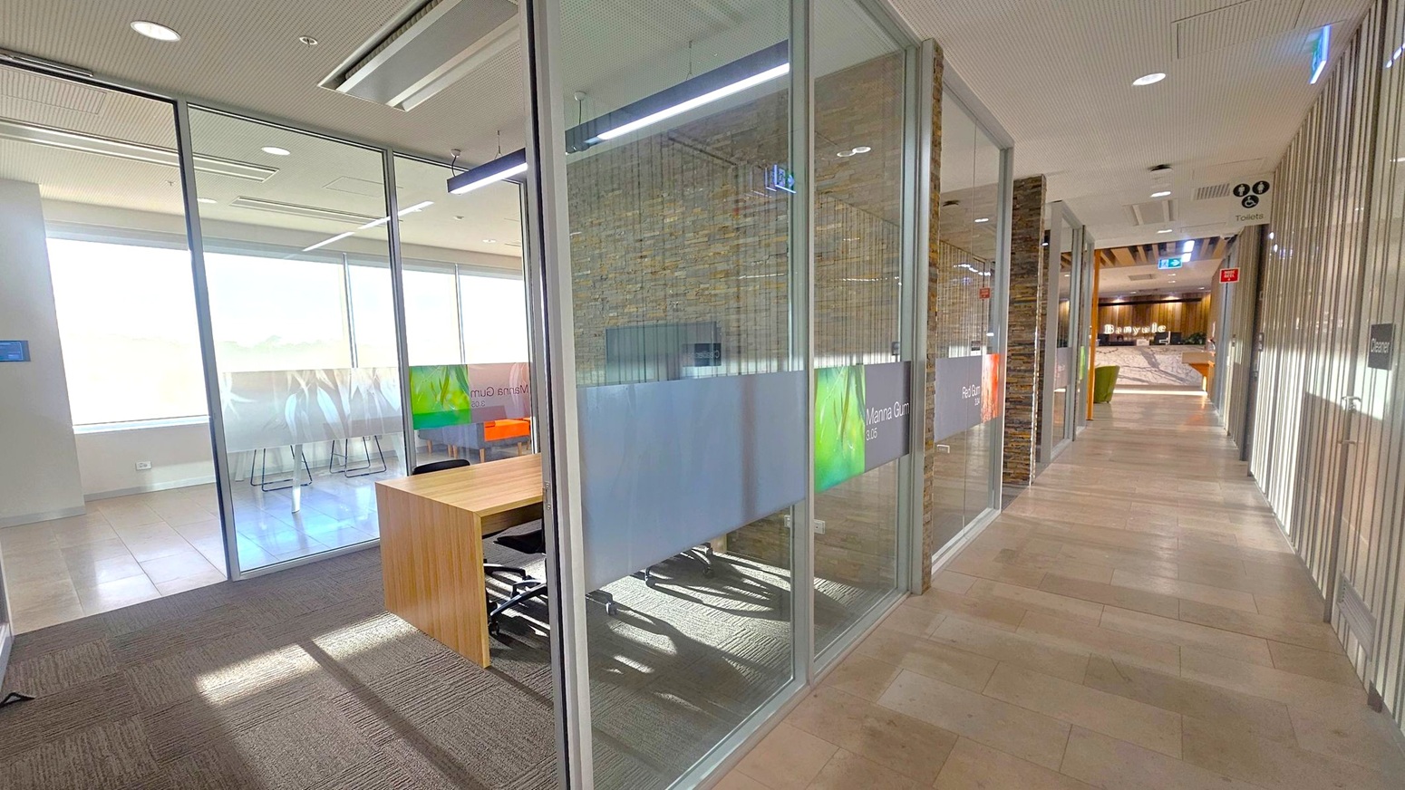 A modern office hallway with glass-walled meeting rooms on the left, featuring frosted glass panels and a wooden desk inside. The floor is a mix of carpet and light-coloured tiles. The ceiling is fitted with recessed lighting and air conditioning vents. On the right, a corridor leads to a reception area with a wooden counter and seating. Signs for toilets and a cleaner's room are visible along the corridor.