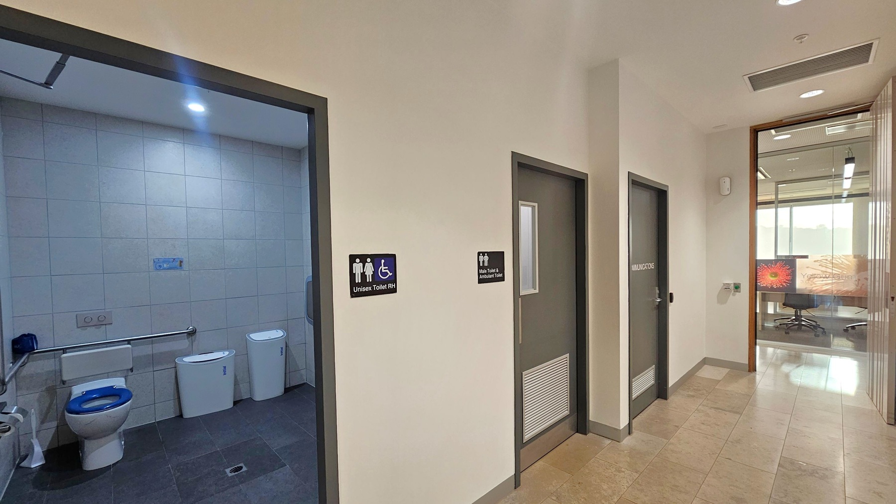 A hallway with three doors on the right. The first door is open, revealing a unisex toilet with accessibility features, including a grab bar and a blue-seated toilet. The second door is labelled "Male Toilet & Ambulant Toilet," and the third door is marked "Communications." On the far right, a glass wall shows an office space with a chair and a colourful design on the glass. The floor is tiled, and the ceiling has recessed lighting and an air vent.