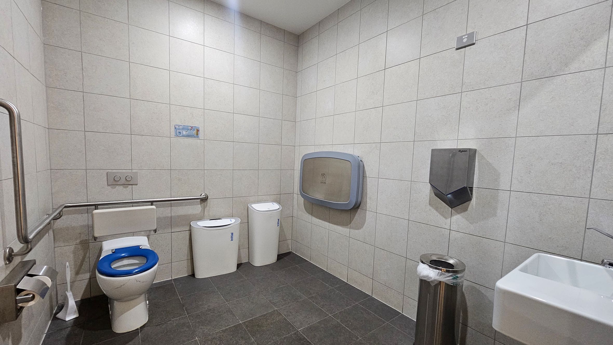 A public restroom with light grey tiled walls and a dark grey tiled floor. On the left, there is a toilet with a blue seat, equipped with a metal grab bar. Next to it are two white bins. On the right wall, a baby changing station and a hand dryer are mounted. Below them, a stainless steel bin with a plastic liner is placed. In the foreground, a white sink with a metal tap is visible. The ceiling is plain and white.