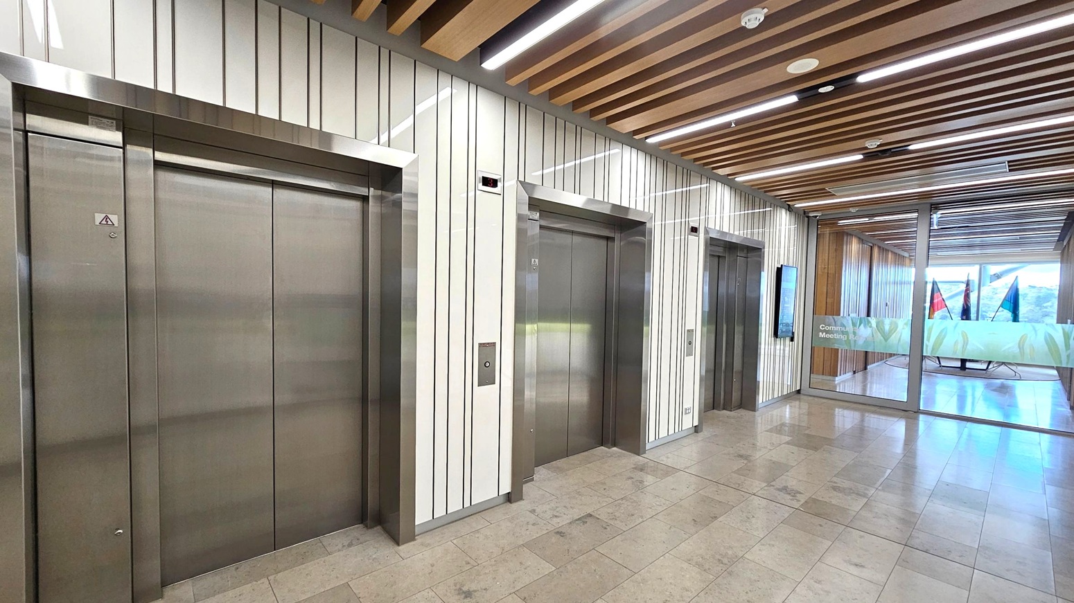 Three stainless steel lift doors are set into a wall with vertical striped panels. The ceiling features wooden slats with integrated lighting. The floor is tiled in a light stone pattern. To the right, glass doors lead to a room with flags and a sign that reads "Community Meeting." The area is well-lit and modern in design.
