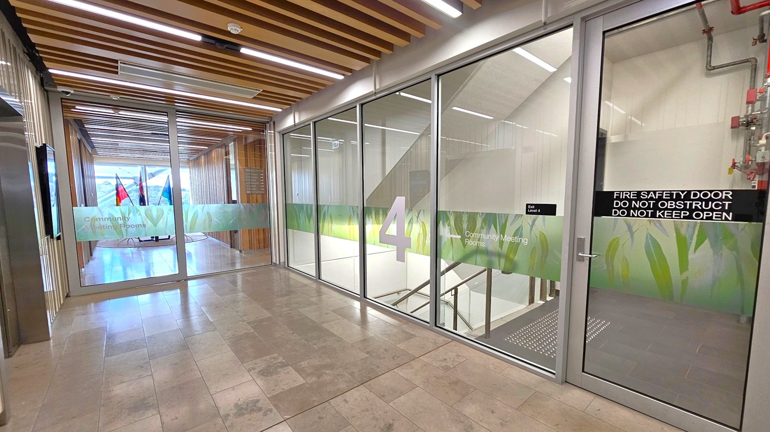 A hallway with a tiled floor and a ceiling featuring wooden slats and recessed lighting. On the right, a glass door marked "Fire Safety Door" leads to a stairwell, with a large number "4" and "Community Meeting Rooms" signage. On the left, a glass wall with a door opens to a corridor with flags visible in the distance. The glass surfaces have a decorative green pattern.