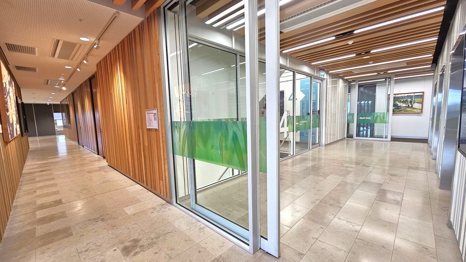 A hallway with light-coloured tiled flooring and a ceiling featuring wooden slats and recessed lighting. On the left, there is a wooden-panelled wall with artwork. To the right, glass doors with green frosted panels lead to a "Staff Only" area. Beyond the doors, a staircase is visible. The walls on the right are white with a framed picture. The corridor extends into the distance, with more artwork visible along the left wall.