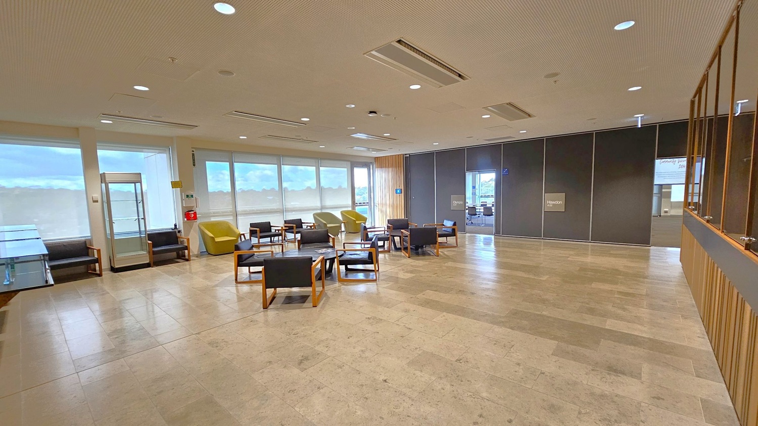 A spacious room with a light-coloured tiled floor and a ceiling with recessed lighting. On the left, there are large windows with blinds, a glass display case, and several black chairs with wooden frames. In the centre, more chairs and small tables are arranged in a seating area. On the right, a dark partition wall with a doorway leads to another room. The far wall has a wooden panel section, and a sign reads "Hawdon."