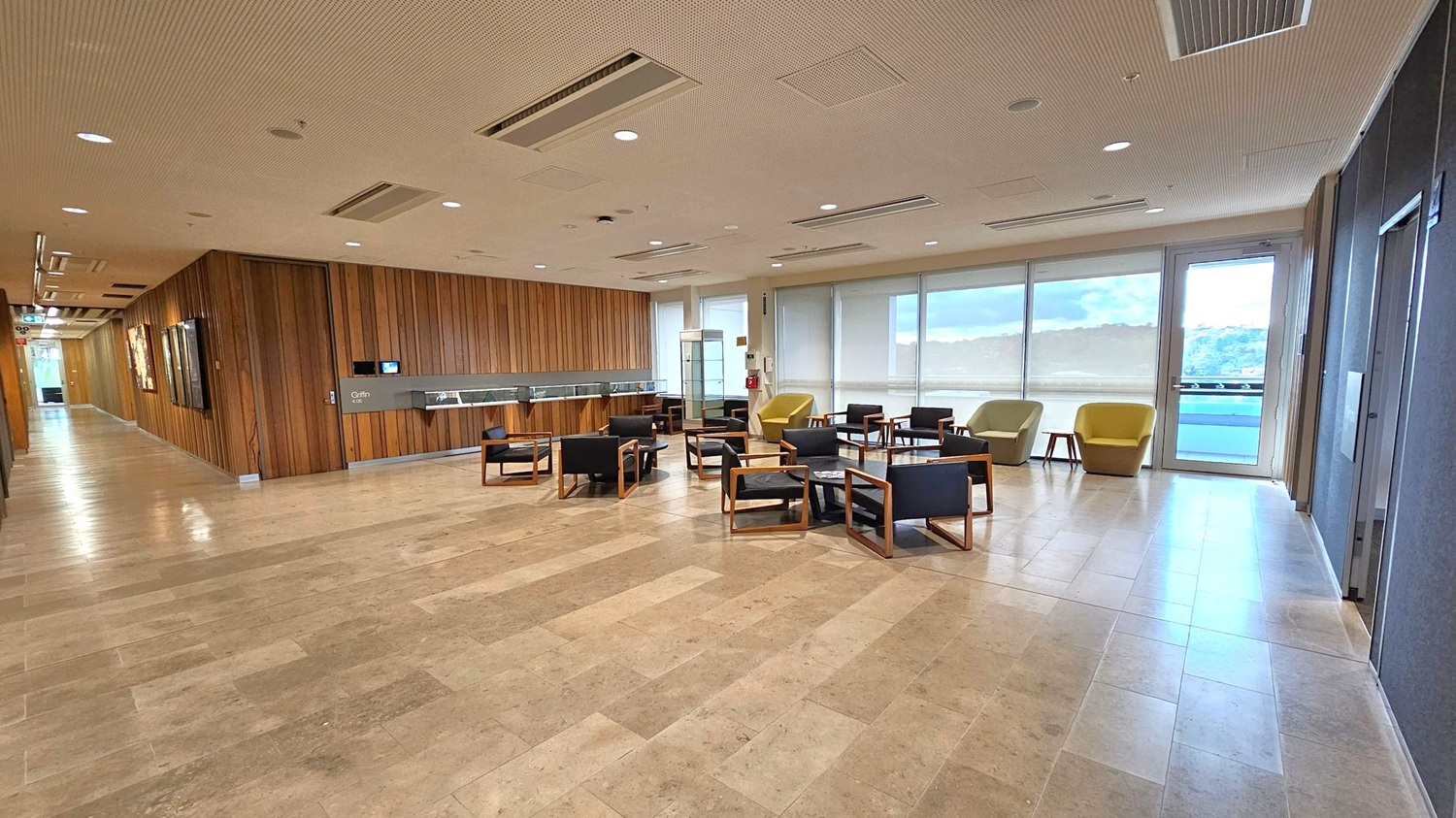 A spacious indoor area with a light-coloured tiled floor and a ceiling with recessed lighting and air vents. On the left, a wooden-panelled wall with artwork and a corridor leading further inside. In the centre, a seating area with black chairs, wooden frames, and small tables. On the right, large windows with blinds and a glass door, offering a view of a landscape outside. Two yellow armchairs are near the windows.