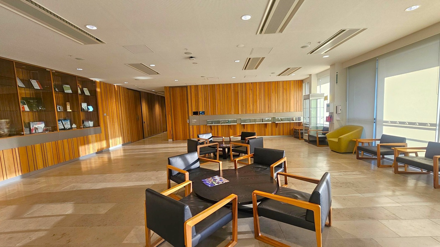 A spacious room with a wooden-panelled wall on the left, featuring display shelves with various items. In the centre, there are several black chairs with wooden frames arranged around circular tables. On the right, a large window with blinds partially drawn allows light to filter in, illuminating a beige armchair. The floor is tiled in a light, neutral colour, and the ceiling is white with recessed lighting.