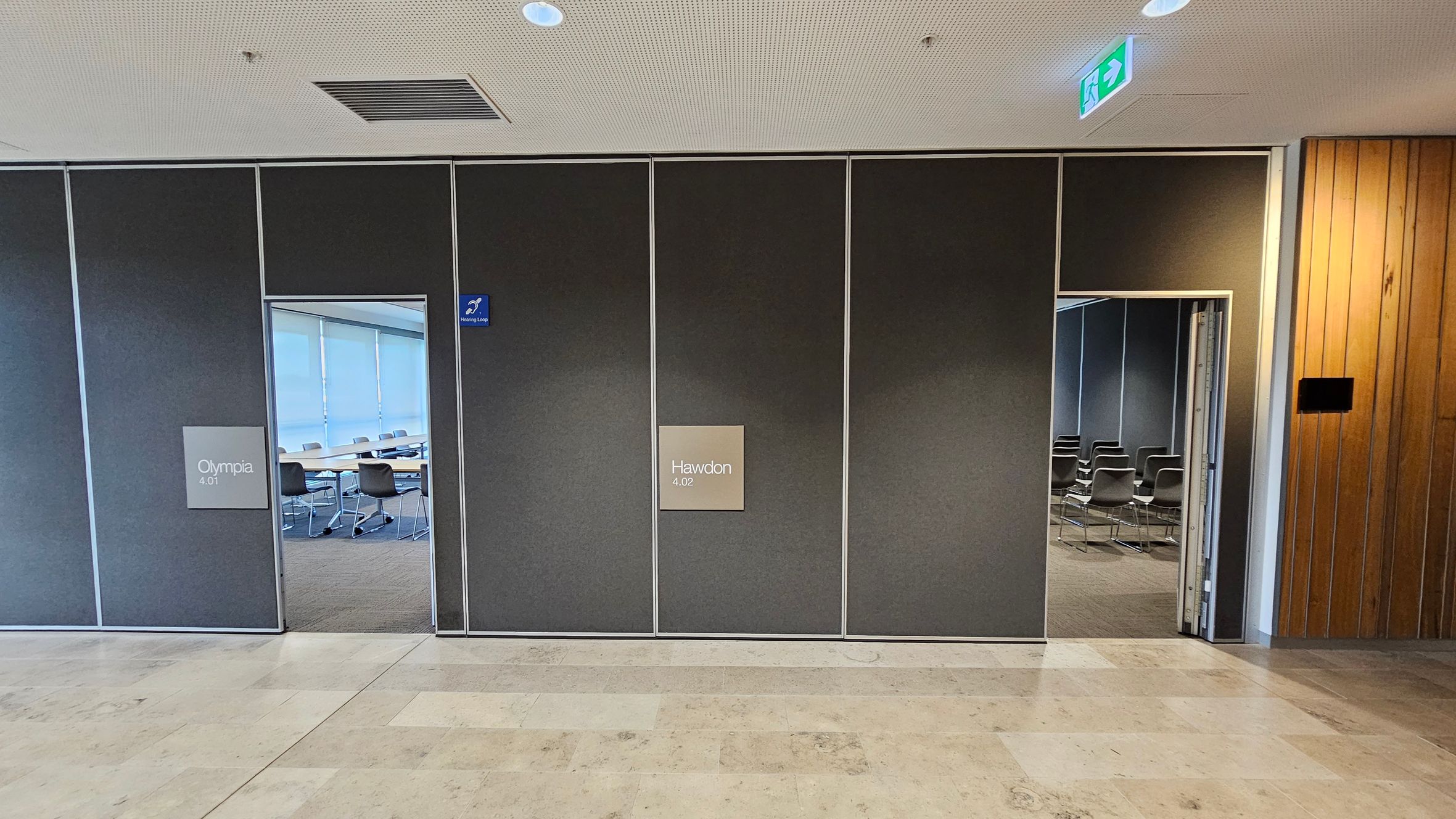 A hallway with two doors leading to separate rooms. On the left, the door labelled "Olympia 4.01" opens to a room with tables and chairs. On the right, the door labelled "Hawdon 4.02" reveals a room with rows of chairs. The wall is dark with a wooden panel on the far right. The ceiling is light-coloured with a vent and recessed lighting. An exit sign is visible above the right door. The floor is tiled in a light, neutral colour.