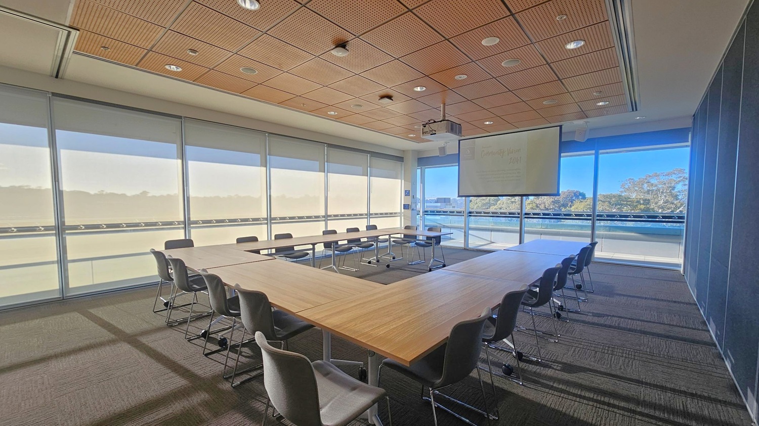 A conference room with a U-shaped arrangement of wooden tables surrounded by grey chairs. The left side features large windows with blinds partially drawn, allowing natural light to enter. On the right, a projector screen hangs from the ceiling, displaying faint text. The background shows a view of trees through the windows. The ceiling is panelled with wood and fitted with recessed lighting. The floor is carpeted in a dark tone.