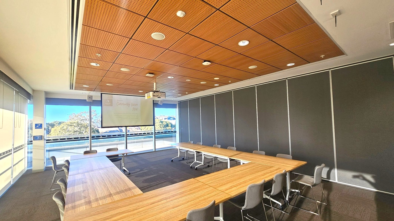 A conference room with a U-shaped arrangement of wooden tables surrounded by grey chairs. The ceiling is wooden with recessed lighting. A projector and screen are mounted at the front, displaying faint text. Large windows on the left offer a view of trees and a building. The floor is carpeted in a dark tone, and the right wall is covered with dark panels.