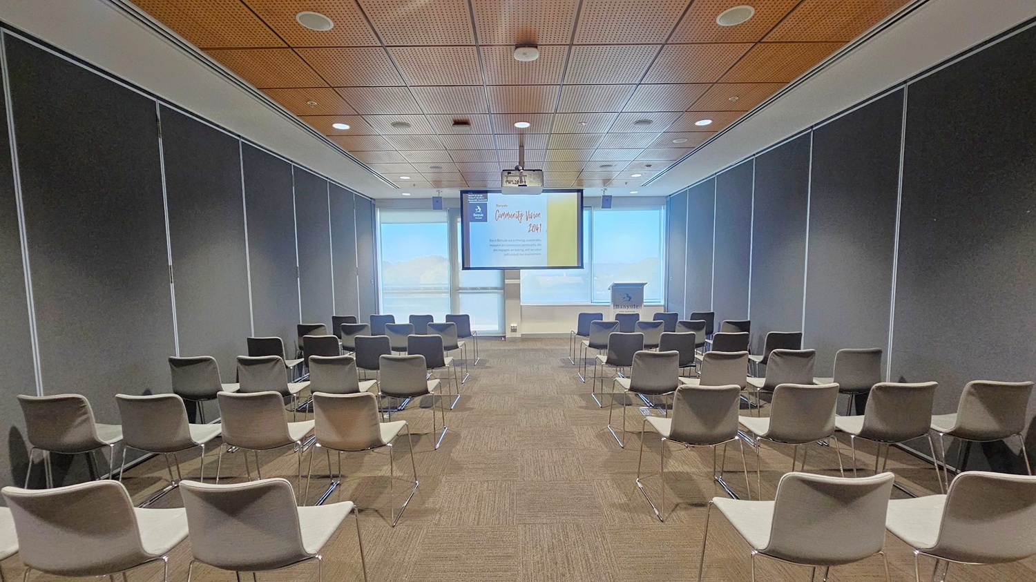 A conference room with rows of grey and beige chairs facing a screen at the front. The screen displays a presentation titled "Community Vision 2041." A podium is positioned to the right of the screen. The walls are lined with dark panels, and the ceiling features wooden panelling with recessed lighting. The floor is carpeted in a brown tone. Large windows at the front allow natural light to filter in, partially covered by blinds.