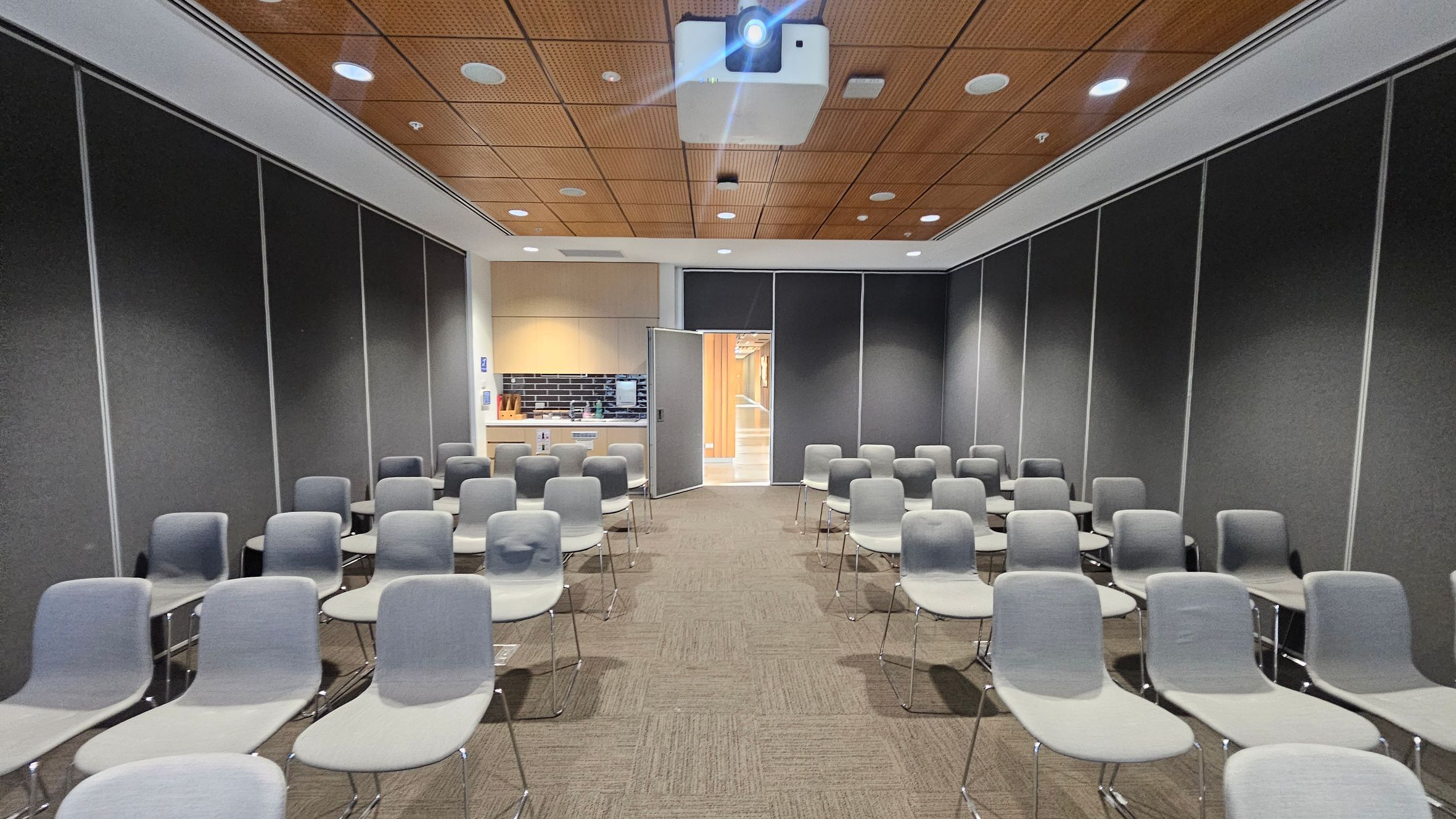 A conference room with rows of grey chairs arranged in a grid pattern. The walls are dark grey with a wooden panelled ceiling featuring recessed lights and a projector. At the back, there's an open door leading to a hallway with wooden accents. To the left of the door, a small kitchenette area with a black tiled backsplash and light wood cabinets is visible. The floor is carpeted in a light brown tone.