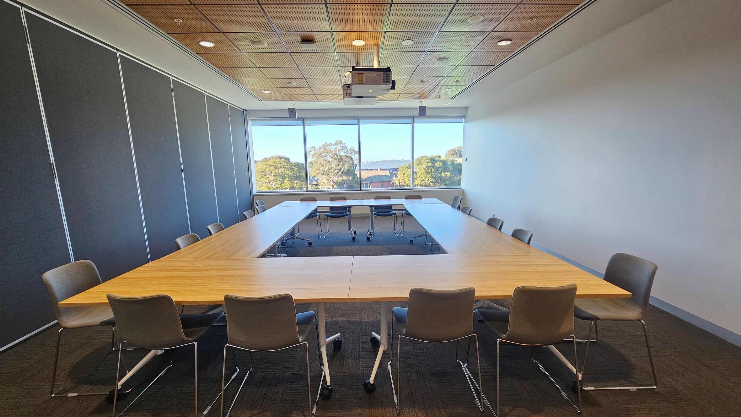 A conference room with a U-shaped wooden table surrounded by grey chairs. The left wall features dark panels, while the right wall is plain white. At the back, large windows offer a view of trees and buildings. The ceiling is wooden with recessed lights and a central projector. The floor is carpeted in a dark colour.