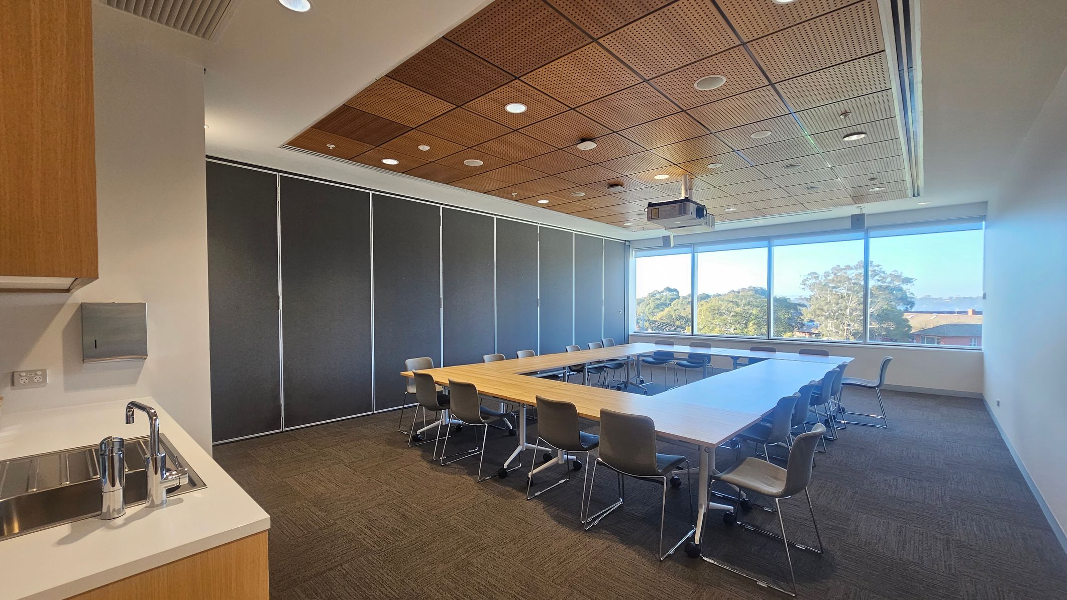 A conference room with a U-shaped arrangement of wooden tables and grey chairs on a carpeted floor. On the left, there's a kitchenette with a sink and a paper towel dispenser. The back wall features large windows with a view of trees and rooftops. The ceiling is wooden with recessed lighting and a projector.