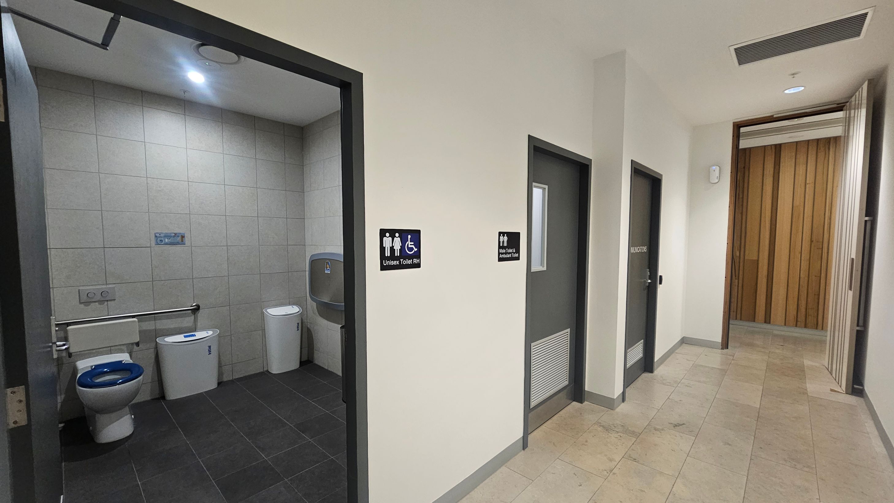 On the left, an open door reveals a unisex toilet with a blue seat, grab bar, and a nappy changing station. The floor is dark grey tile. In the centre, a sign indicates "Unisex Toilet RH" next to a closed door marked "Male Toilet & Ambulant Toilet." The corridor on the right has light-coloured tiles and leads to a wooden panelled area. The ceiling is plain with recessed lighting.