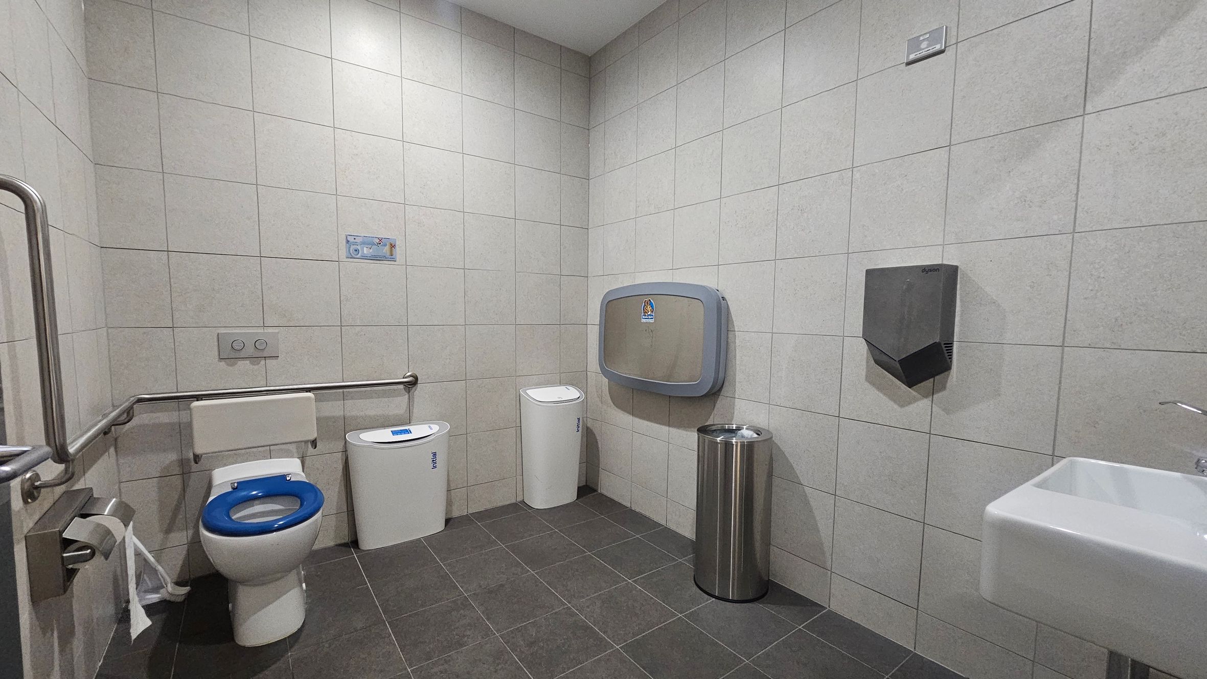 A public restroom with light grey tiled walls and a dark grey tiled floor. On the left, a toilet with a blue seat and a grab bar beside it. A toilet paper holder is mounted on the wall. In the centre, a nappy changing station is attached to the wall above a white bin. On the right, a stainless steel bin and a hand dryer are mounted on the wall. A white sink with a silver tap is visible in the foreground. The ceiling is white with recessed lighting.