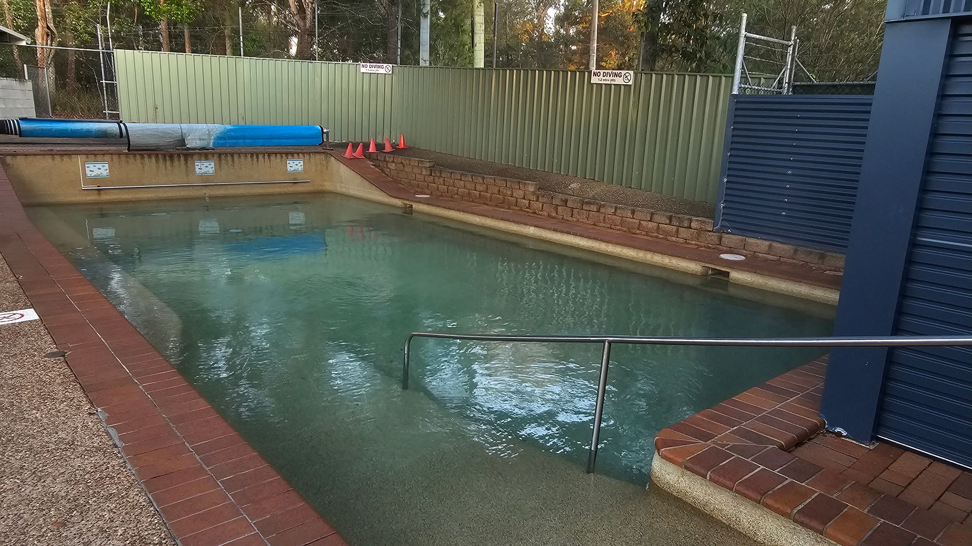An outdoor swimming pool with clear water is surrounded by a brick and concrete deck. On the left, there are orange traffic cones and a blue pool cover. A green metal fence runs along the back, with trees visible behind it. On the right, a building with a corrugated metal roof and a light-coloured brick wall has signs and a gate leading inside. A "No Diving" sign is attached to the fence. The area is shaded by a large canopy overhead.
