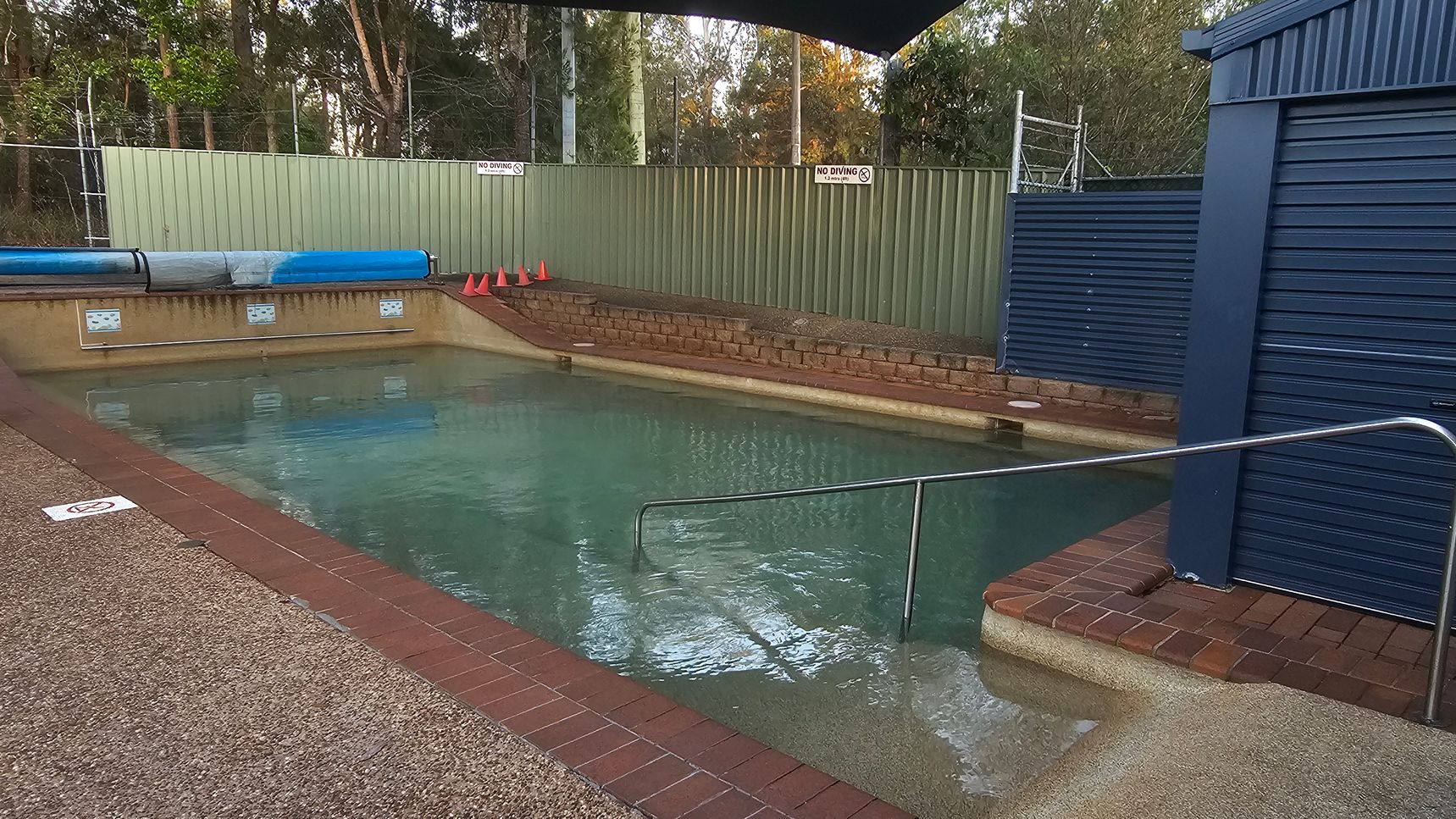 An outdoor swimming pool with clear water is surrounded by a brick edge. On the right, there's a blue metal shed with a roller door. A metal handrail leads into the pool. In the background, a green corrugated metal fence with "No Diving" signs is visible, along with a row of orange traffic cones. Behind the fence, there are tall trees. The pool deck is made of textured concrete.