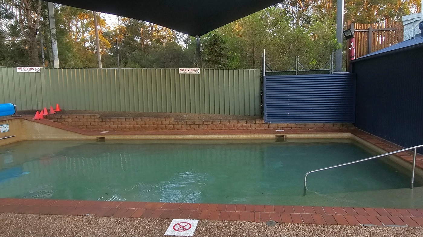 A small swimming pool with clear water is surrounded by a brick and concrete deck. On the left, there are orange traffic cones and a "No Diving" sign on a green metal fence. The right side features a blue corrugated metal wall and a metal handrail leading into the pool. Trees and foliage are visible in the background, and a dark canopy provides shade over the pool area.