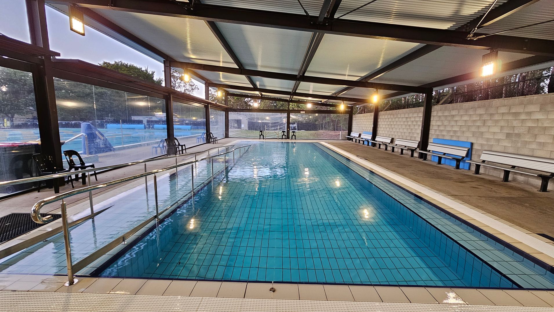 An indoor swimming pool with clear blue water, surrounded by a metal railing on the left. The pool is enclosed by transparent walls, allowing a view of the outdoor area. On the right, there are several white benches against a grey brick wall. The ceiling is made of metal panels with lights hanging from it. The floor around the pool is tiled.
