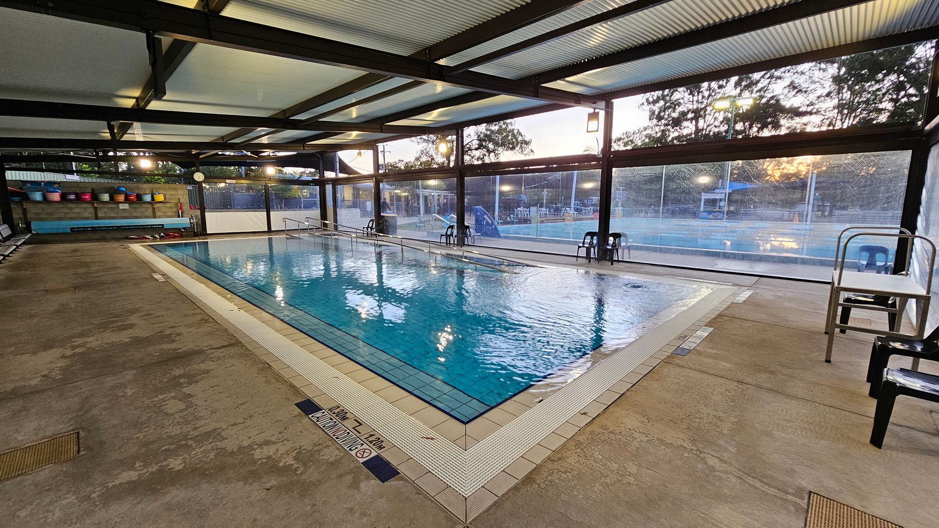 Indoor swimming pool with clear water, surrounded by a tiled edge. On the left, there are colourful buckets on a shelf and a bench. The floor is concrete with some discolouration. Large windows on the right and background provide a view of an outdoor area with trees and a slide. Several black chairs are placed around the pool. The ceiling has exposed beams and corrugated panels.