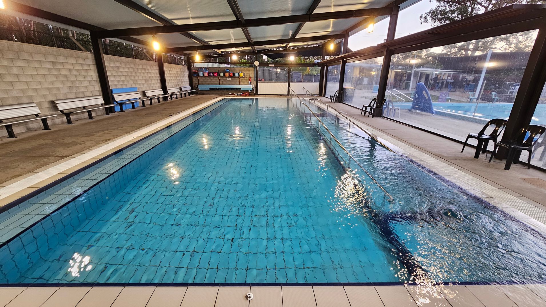 An indoor swimming pool with clear blue water, surrounded by a tiled deck. On the left, there are several white benches against a grey brick wall. On the right, black plastic chairs are placed along a glass wall, offering a view of an outdoor area with trees. The ceiling is supported by black beams, and overhead lights illuminate the space. At the far end, colourful buckets are stacked on a shelf.