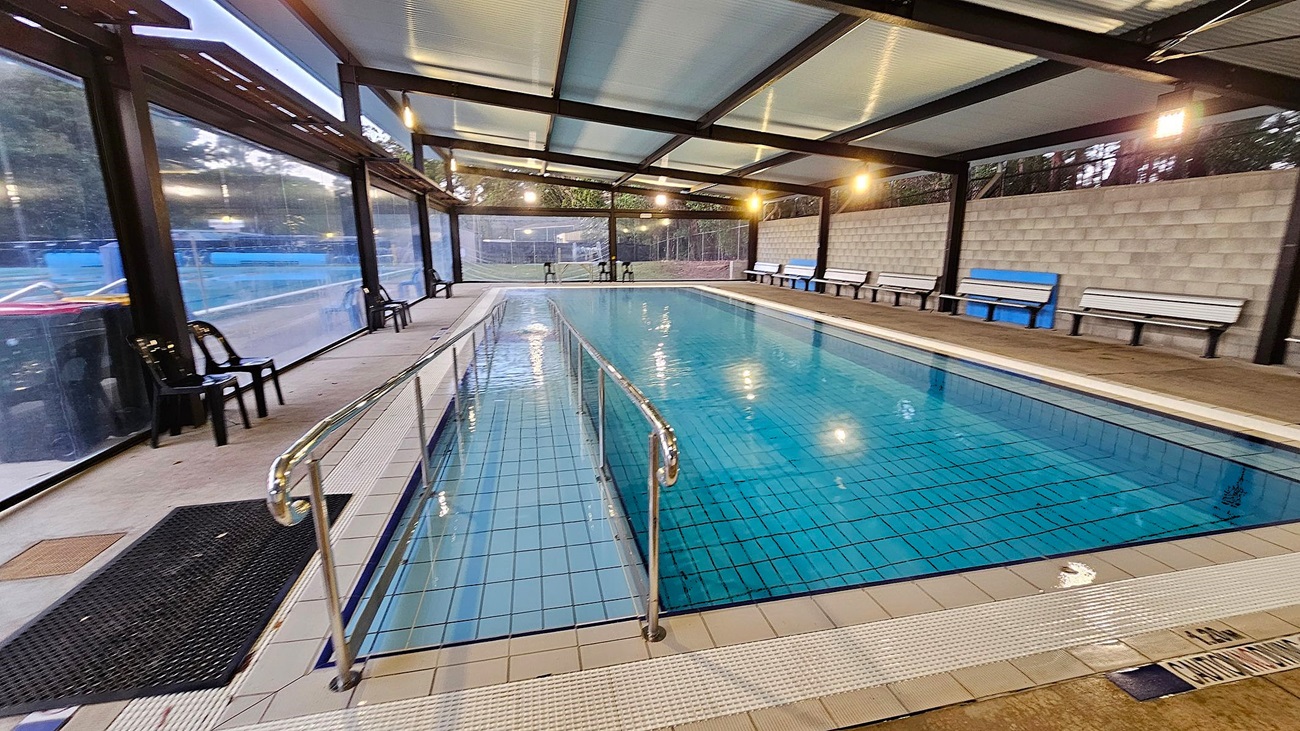 An indoor swimming pool with a metal handrail running down the centre. The pool is surrounded by a tiled deck. On the left, there are black plastic chairs and large glass windows. On the right, a concrete block wall with white benches and a blue bench. The ceiling is made of translucent panels with exposed beams, and the floor is tiled. The area is well-lit with overhead lights.