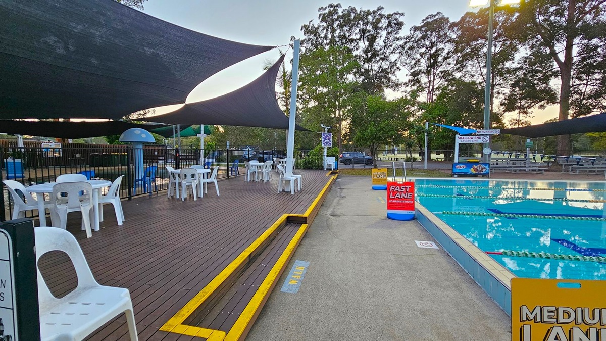 On the left, there is a wooden deck with white plastic tables and chairs under large black shade sails. A blue mushroom-shaped water feature is visible near a fenced area. On the right, a swimming pool with lane markers is labelled "FAST LANE" and "MEDIUM LANE". Signs indicate "NO RUNNING" and "SHALLOW END". Trees and a car park are in the background, with picnic tables scattered around. The ground is a mix of wood and concrete.