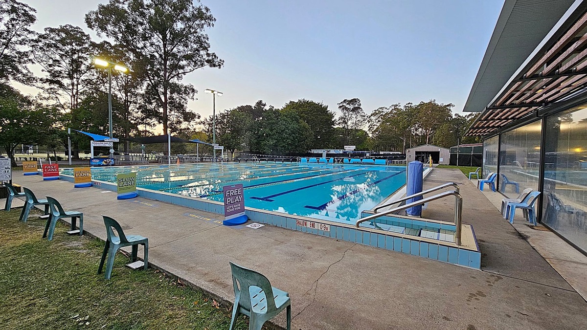 Outdoor swimming pool with lane markers, surrounded by signs indicating lane speeds such as "Fast Lane" and "Slow Lane". On the left, green plastic chairs are placed on a grassy area. On the right, a building with a glass facade and blue chairs. Trees and tall lights are in the background. The pool deck is concrete, and the water is calm.